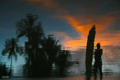 Fotografia que mostra os reflexos produzidos pela vazante do mar na areia. Os tons, muito característicos, são provenientes da hora dourada - o pôr do sol - momento mágico onde o dia vira noite, onde o dia vira lembrança. Aqui um homem está com seu long board, provavelmente indo descansar após horas no mar.